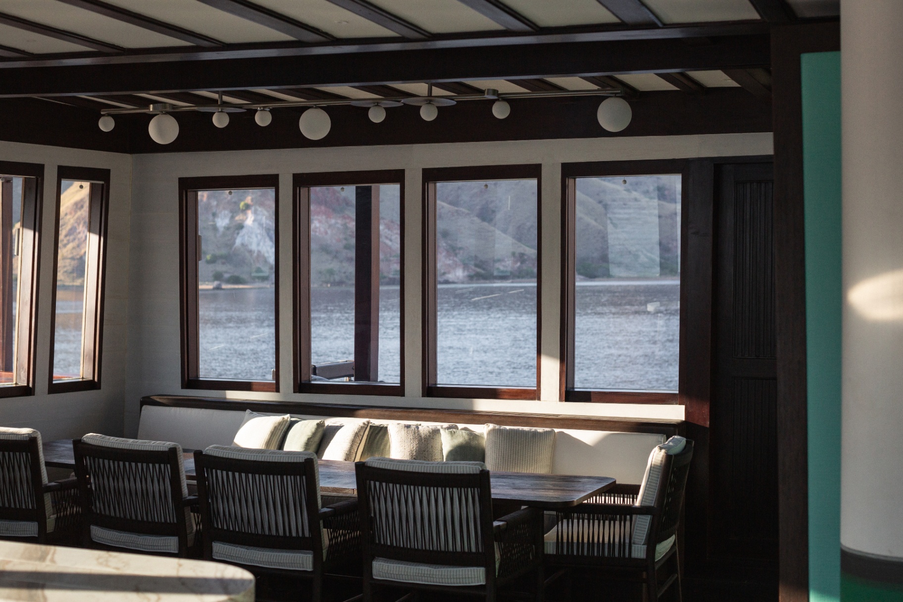 Indoor dining banquette in Vela's saloon with rattan chairs and floor-to-ceiling windows onto Komodo cliffs