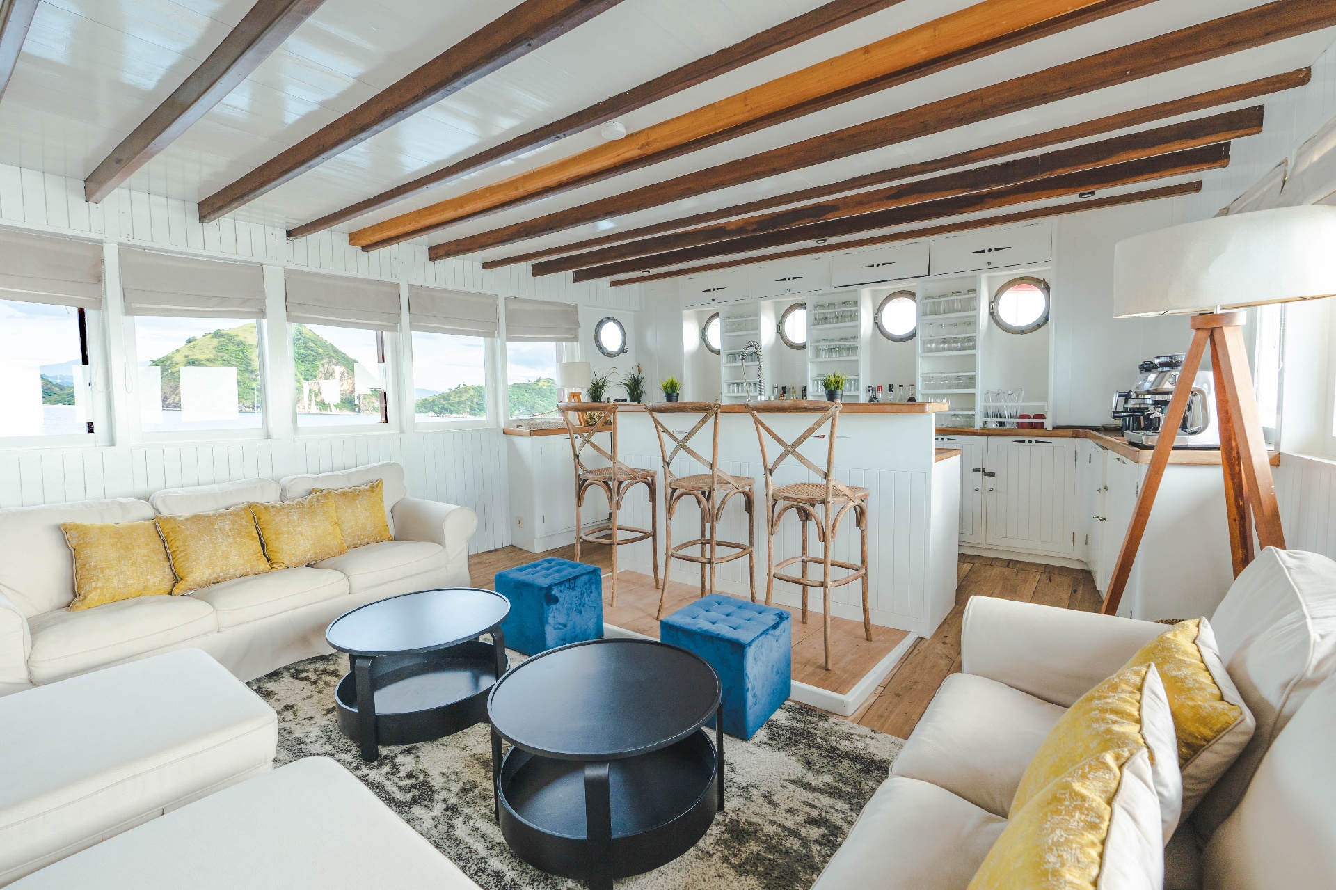 Mischief indoor saloon with white sofas, yellow cushions, coffee tables, bar with rattan stools and Komodo island views through the windows