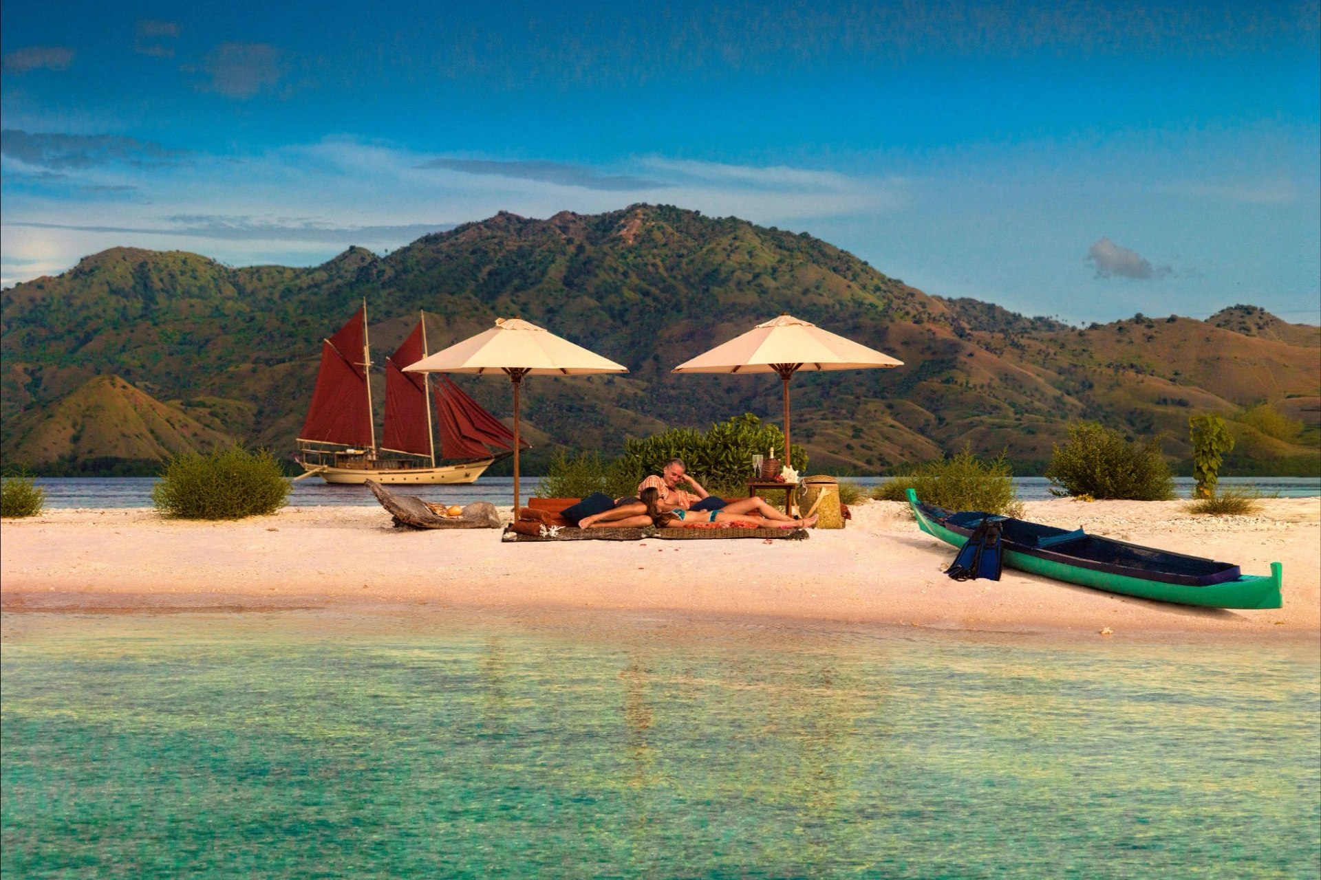 Si Datu Bua anchored offshore while guests relax on a white-sand beach under twin umbrellas with a dugout canoe
