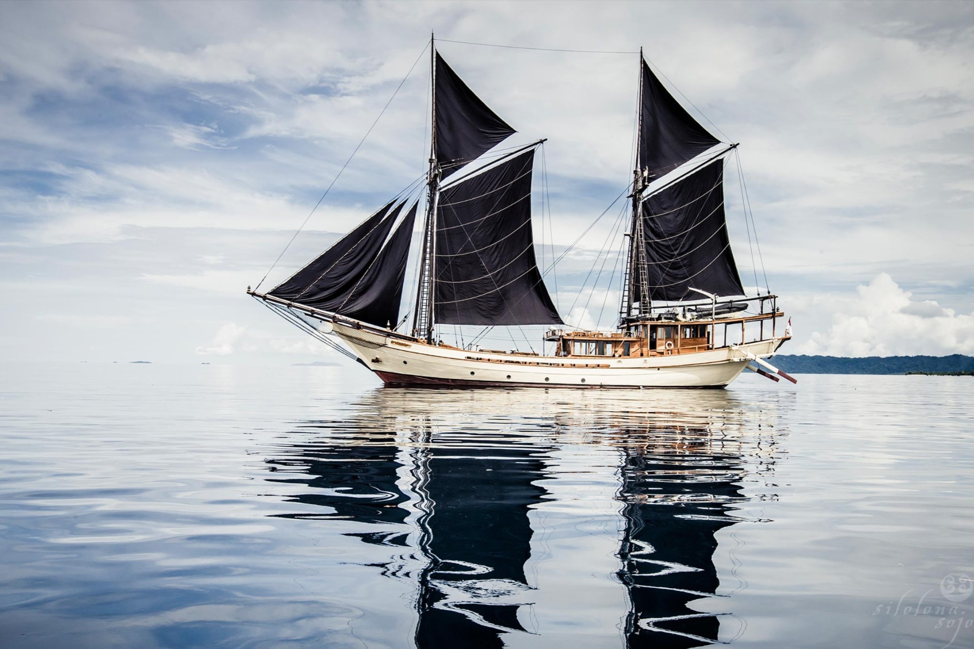 Silolona on mirror-calm water with black sails reflected below