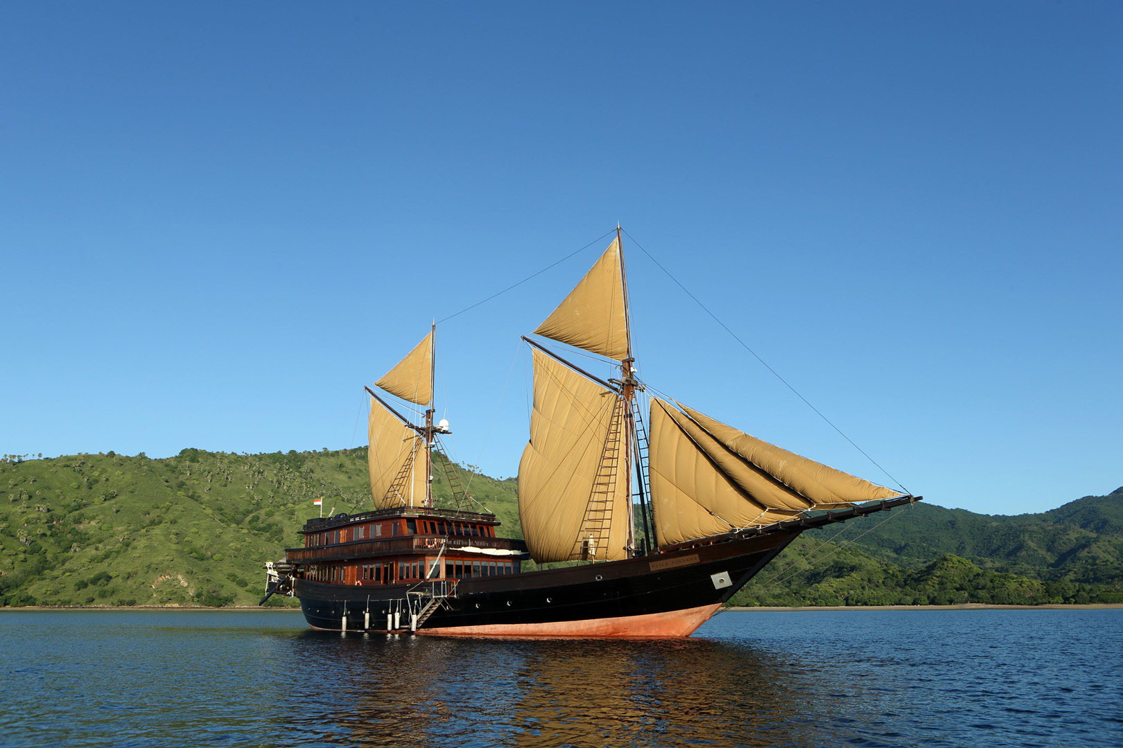 Alila Purnama luxury phinisi under full sail in a green Indonesian bay — Raja Ampat private yacht charter