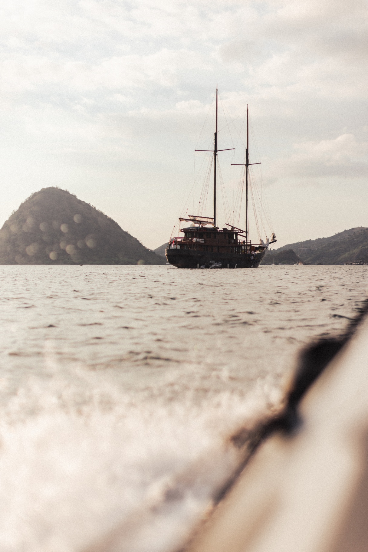 Vela anchored off Labuan Bajo with twin masts silhouetted against soft afternoon light and hills behind