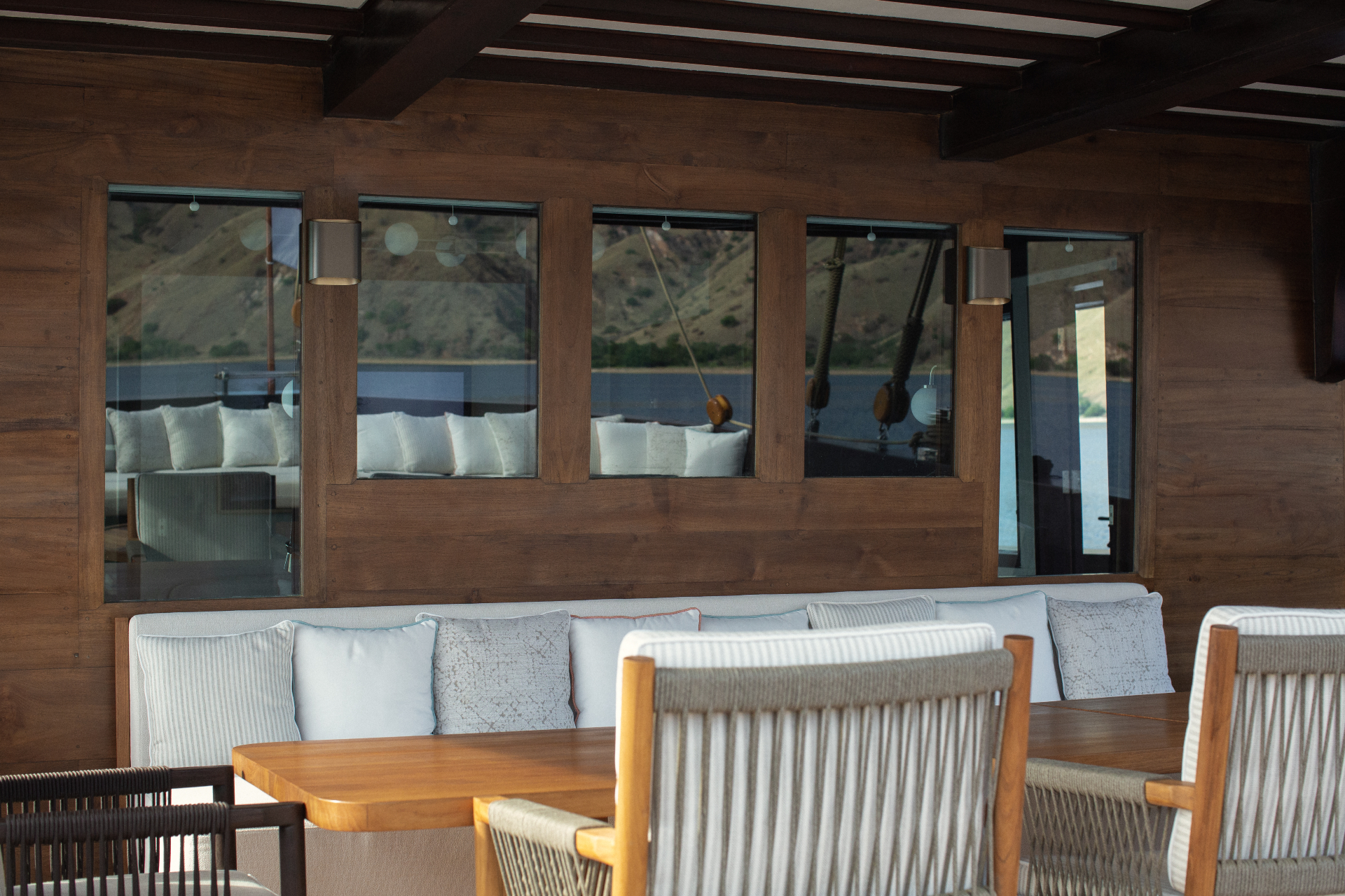 Main deck outdoor dining banquette in warm teak with picture windows reflecting arid Komodo hills