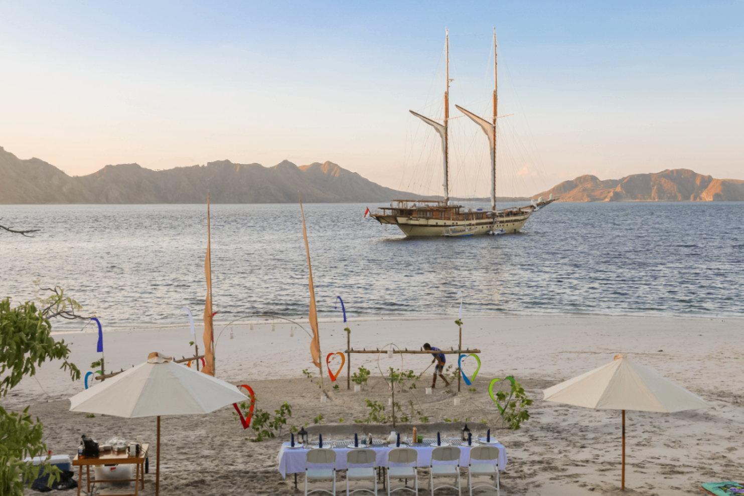 Long beach dinner table set on white sand at sunset with Lamima anchored offshore in front of Komodo hills