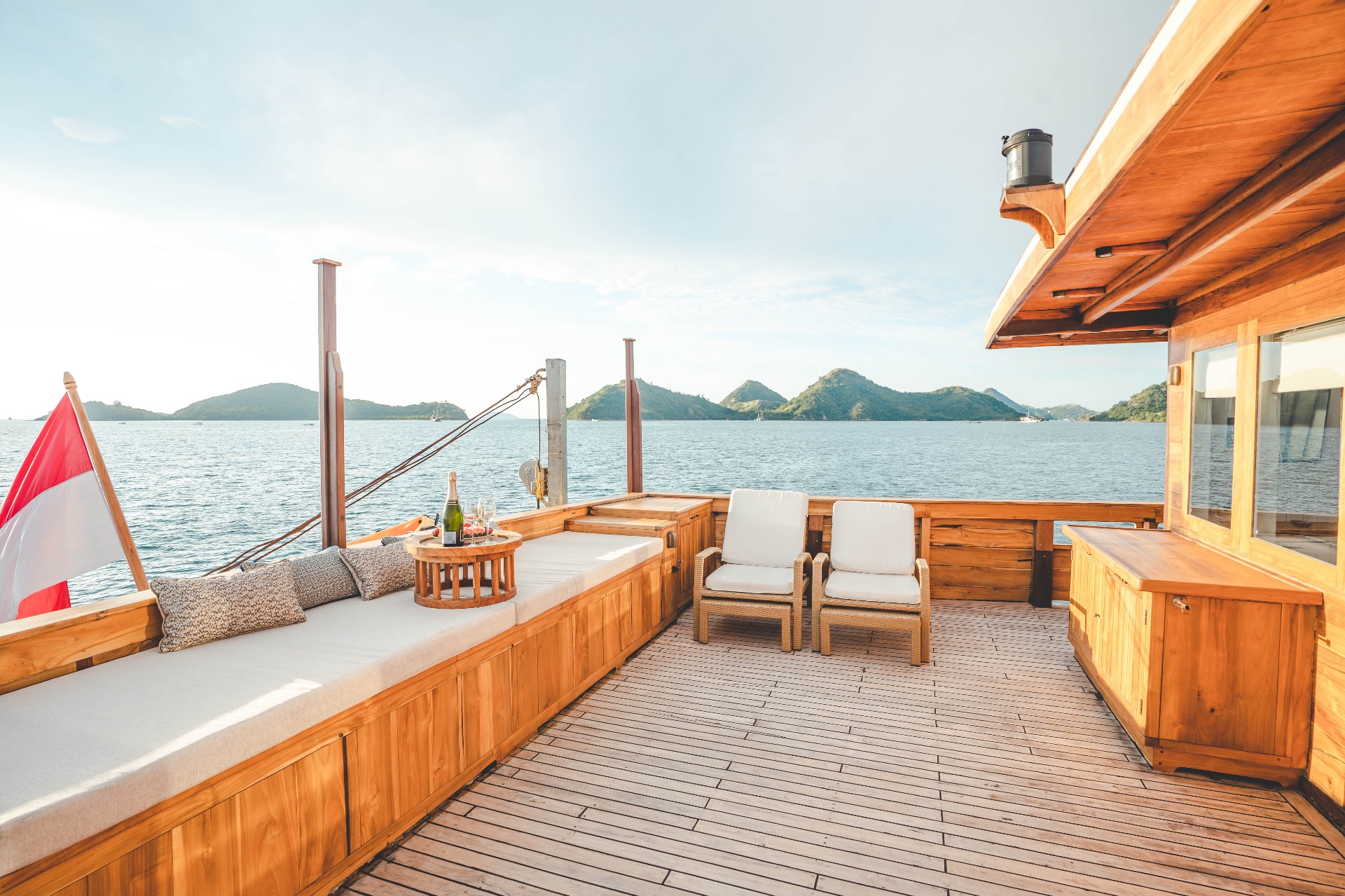 Mischief foredeck lounge with long cushioned bench, rattan chairs and Indonesian flag against island horizon
