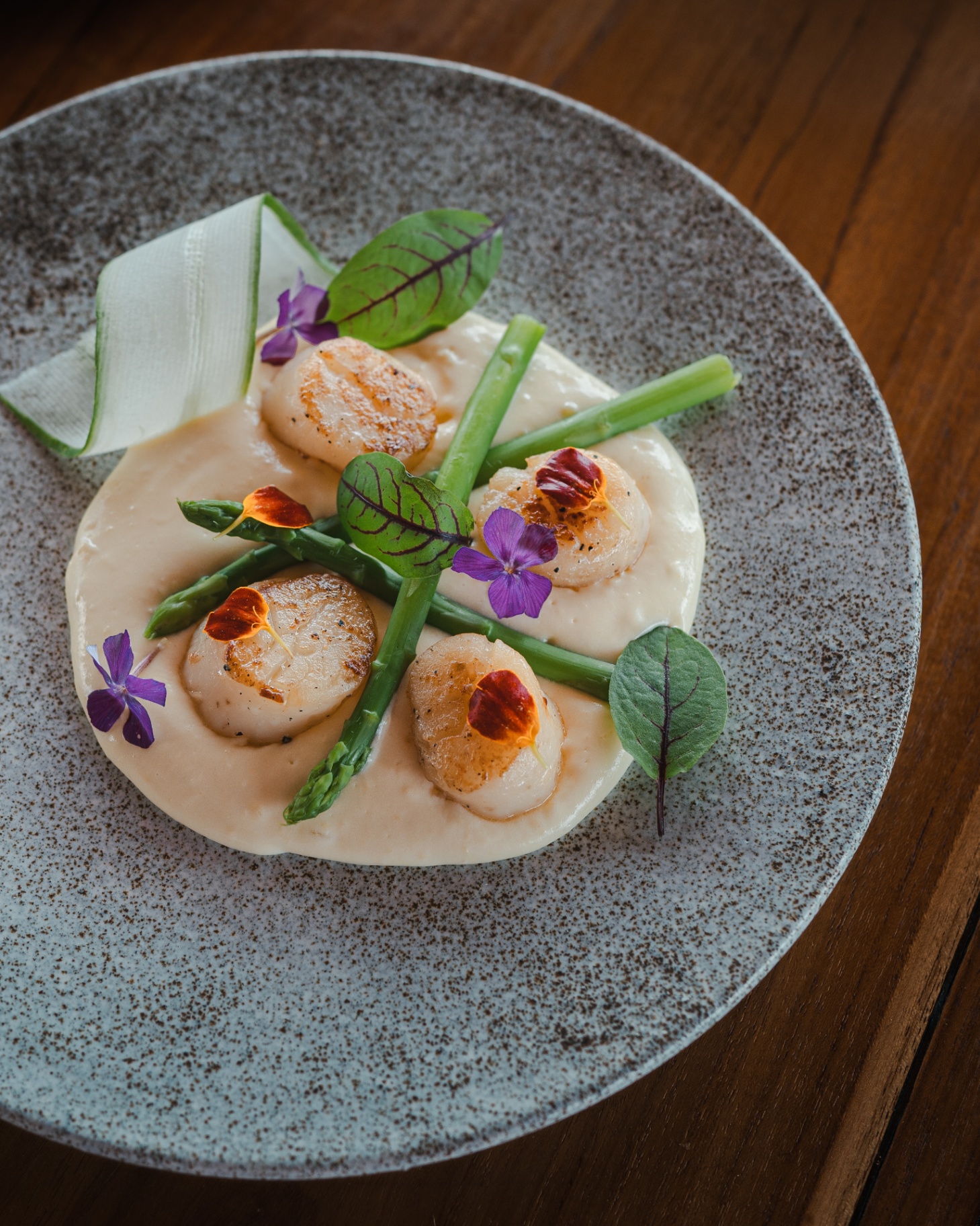 Seared scallops with asparagus, cream sauce and edible flowers plated on stoneware aboard Mischief