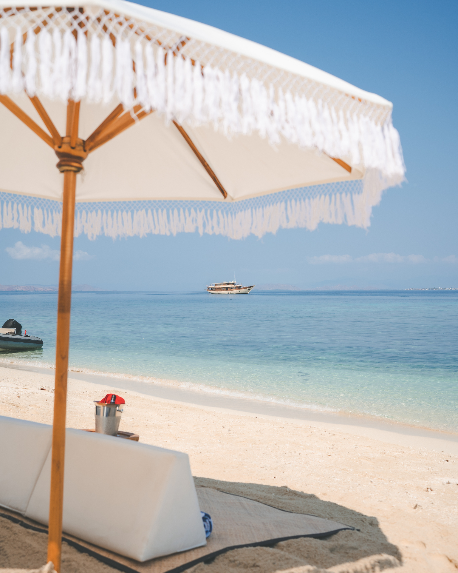 View from beneath a fringed beach umbrella looking out to Mischief anchored offshore in calm blue water