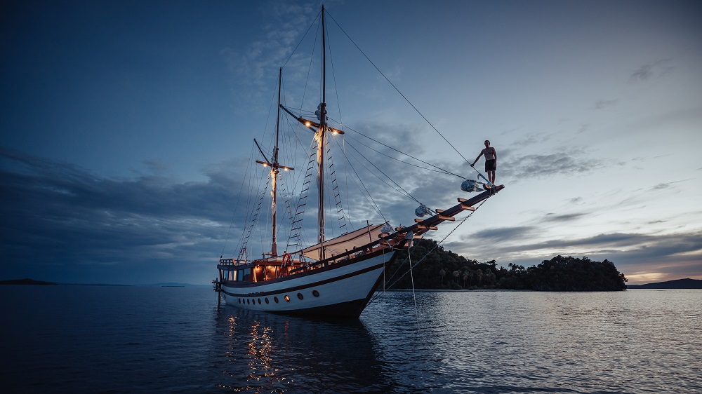 Guest standing on Senja's bowsprit at dusk with the lit phinisi behind on calm water