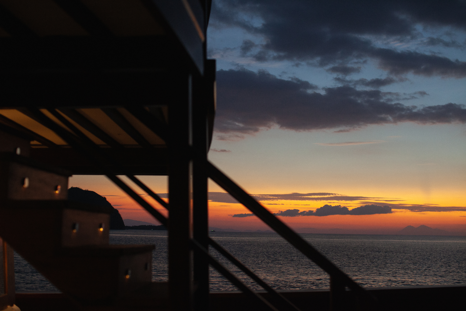 Sunset view from Vela's deck with silhouetted rigging, an island headland and an orange horizon