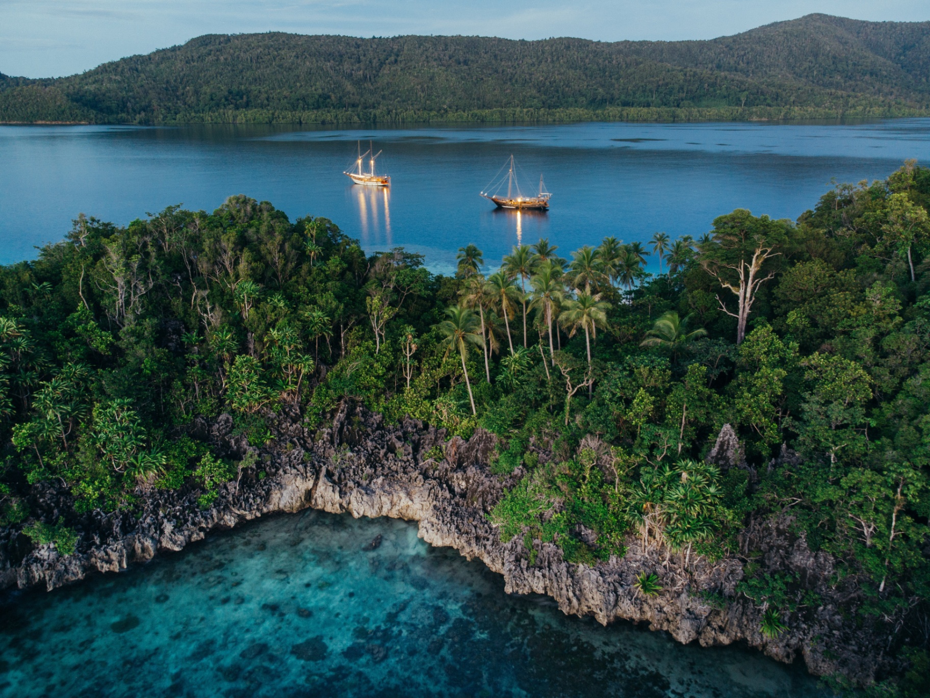 Senja and a sister phinisi lit up at dusk in a palm-fringed Raja Ampat lagoon