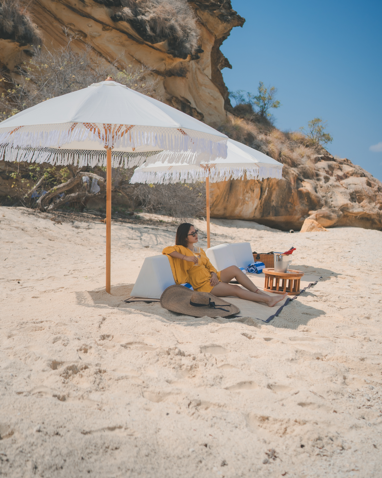 Guest in a yellow dress relaxing with a glass of wine under a fringed white umbrella on a private Komodo beach set up by Mischief's crew