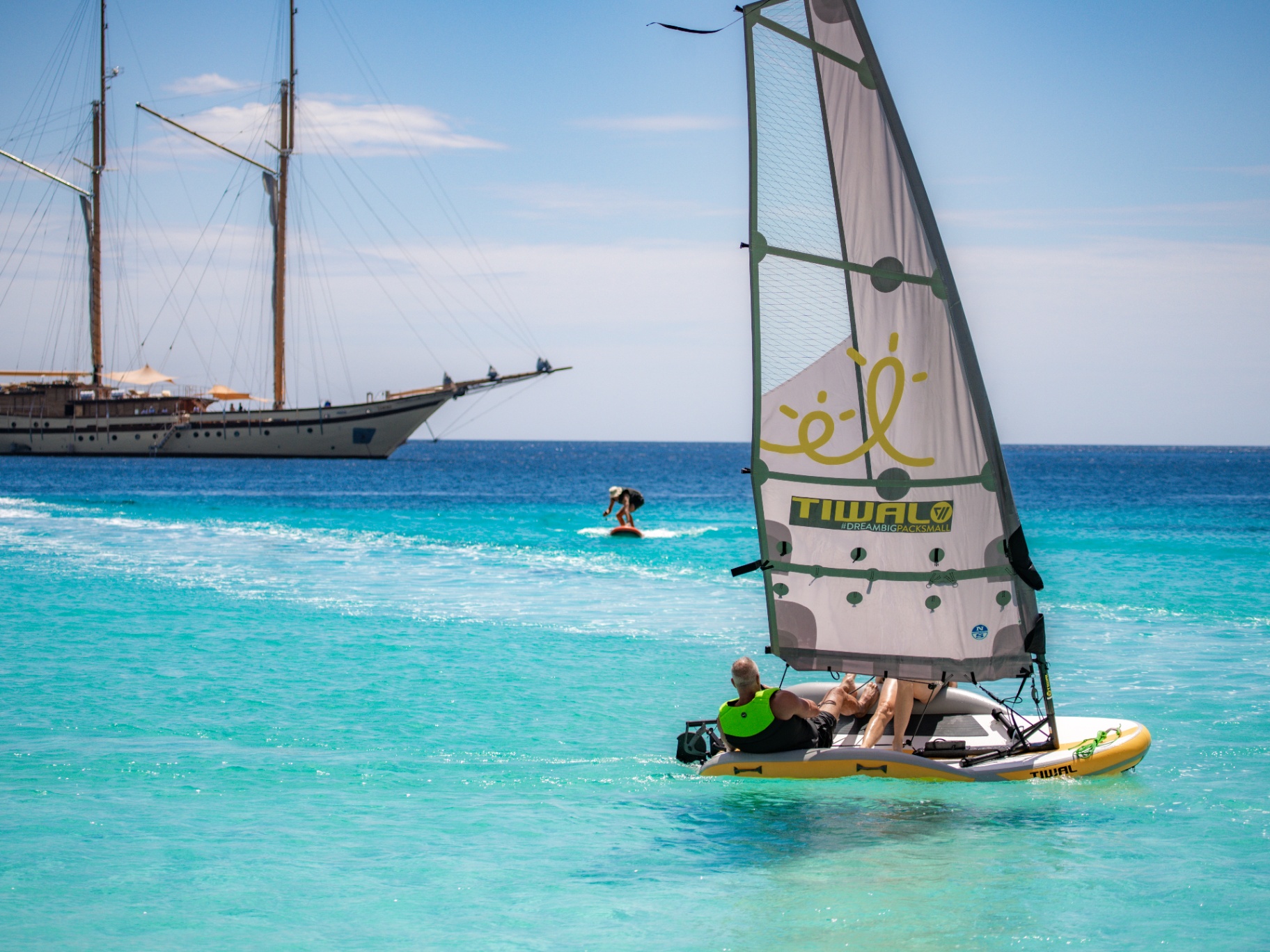 Guest sailing a Tiwal dinghy on bright turquoise water with Lamima anchored offshore and a wing-foiler in the distance