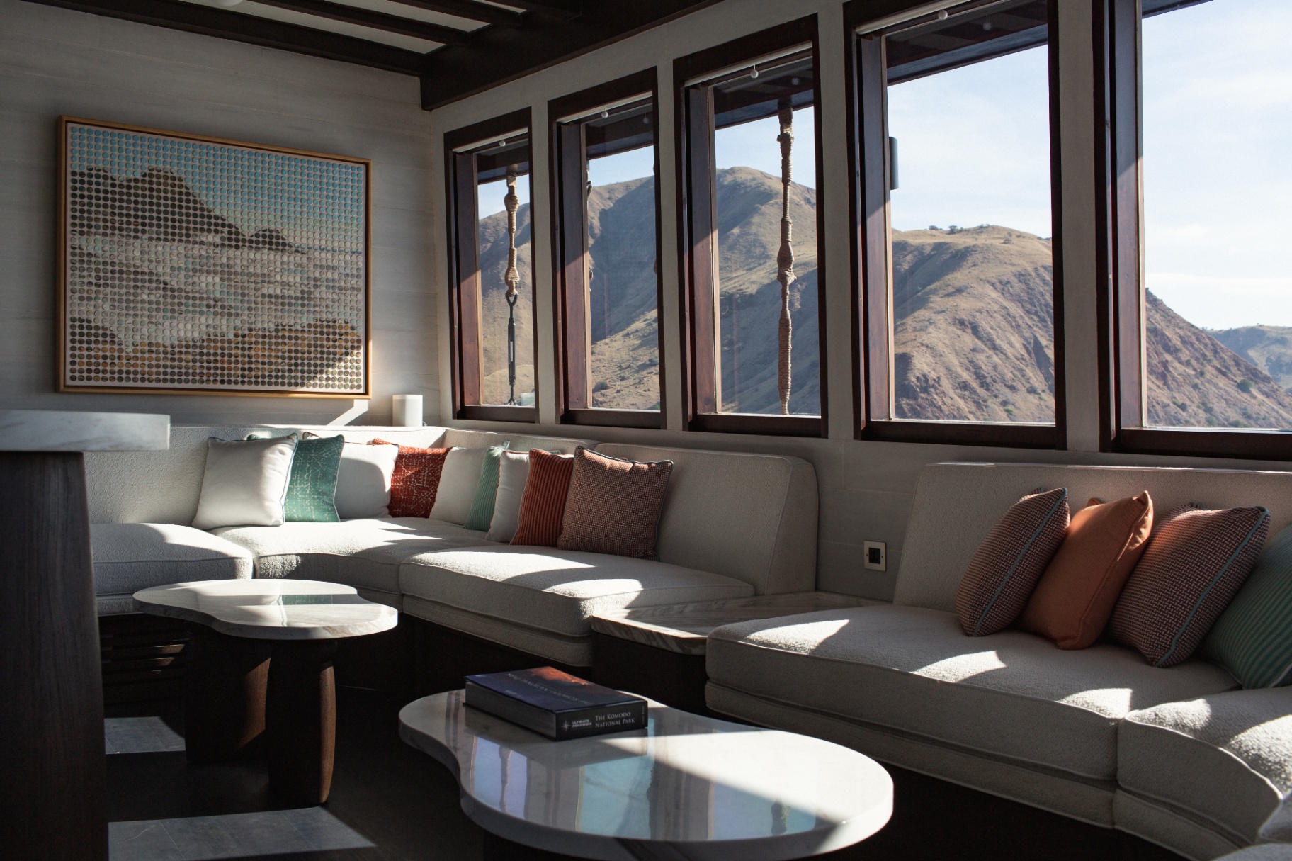 Main saloon lounge on Vela with L-shaped sofa, marble tables and picture windows framing arid Komodo hills
