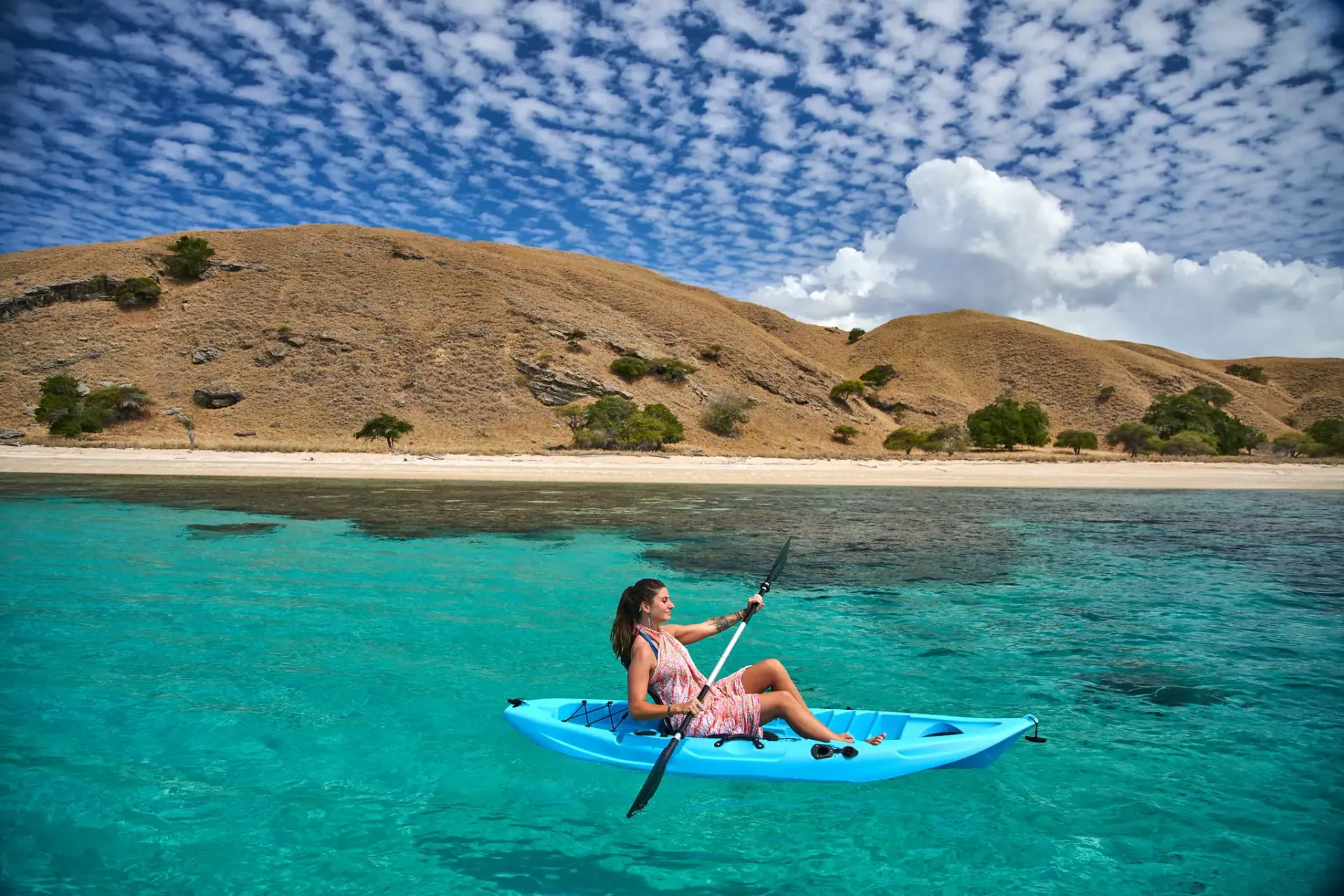 Guest kayaking in turquoise water off a Komodo beach with hills behind
