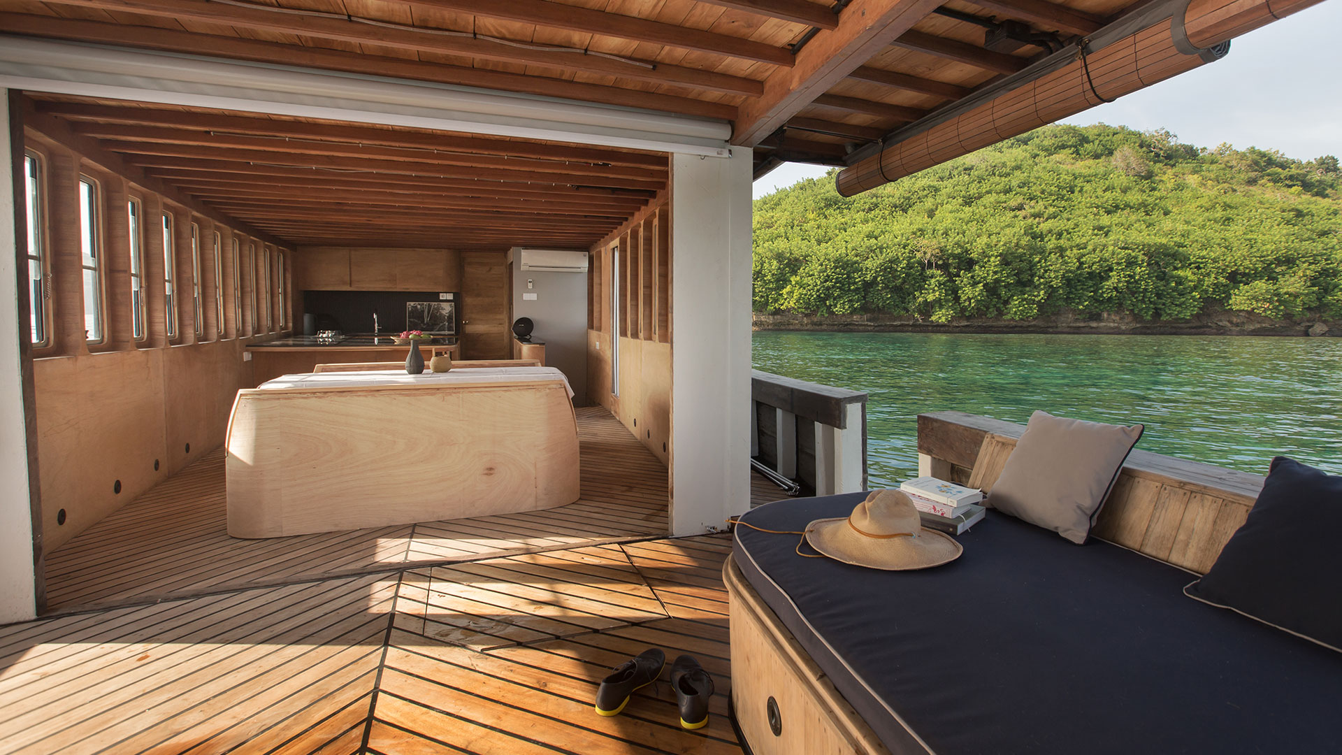 Splendour's open-plan interior with dining area, galley, teak flooring and lounge cushions overlooking a green shoreline
