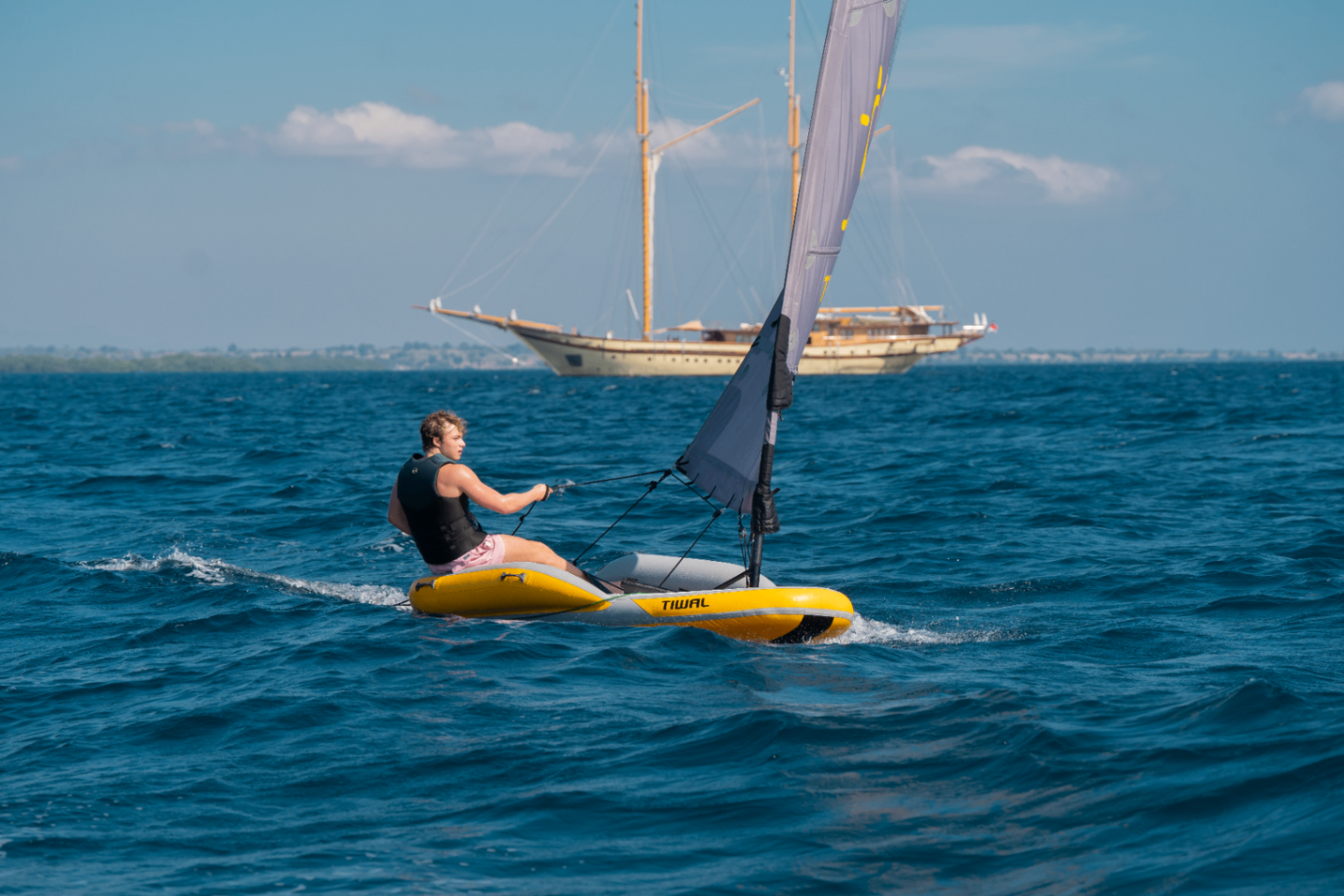Young guest sailing a Tiwal inflatable dinghy on deep blue open water with Lamima under bare poles in the background