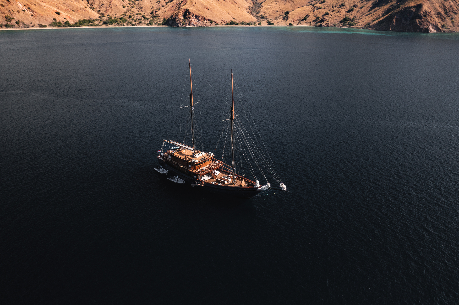 Aerial view of Vela at anchor in a sheltered Komodo bay with tenders alongside and arid hills behind