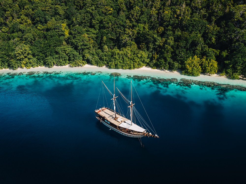 Aerial of Senja anchored on deep blue water beside a white sand beach and coral reef in Raja Ampat