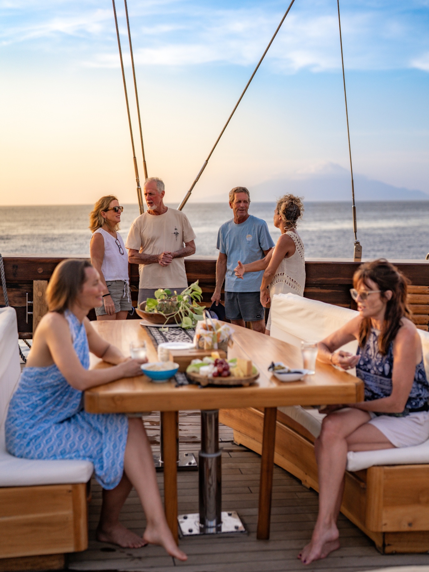 Group of guests enjoying sunset cocktails and cheese board on Lamima's aft deck with the sun setting over the sea
