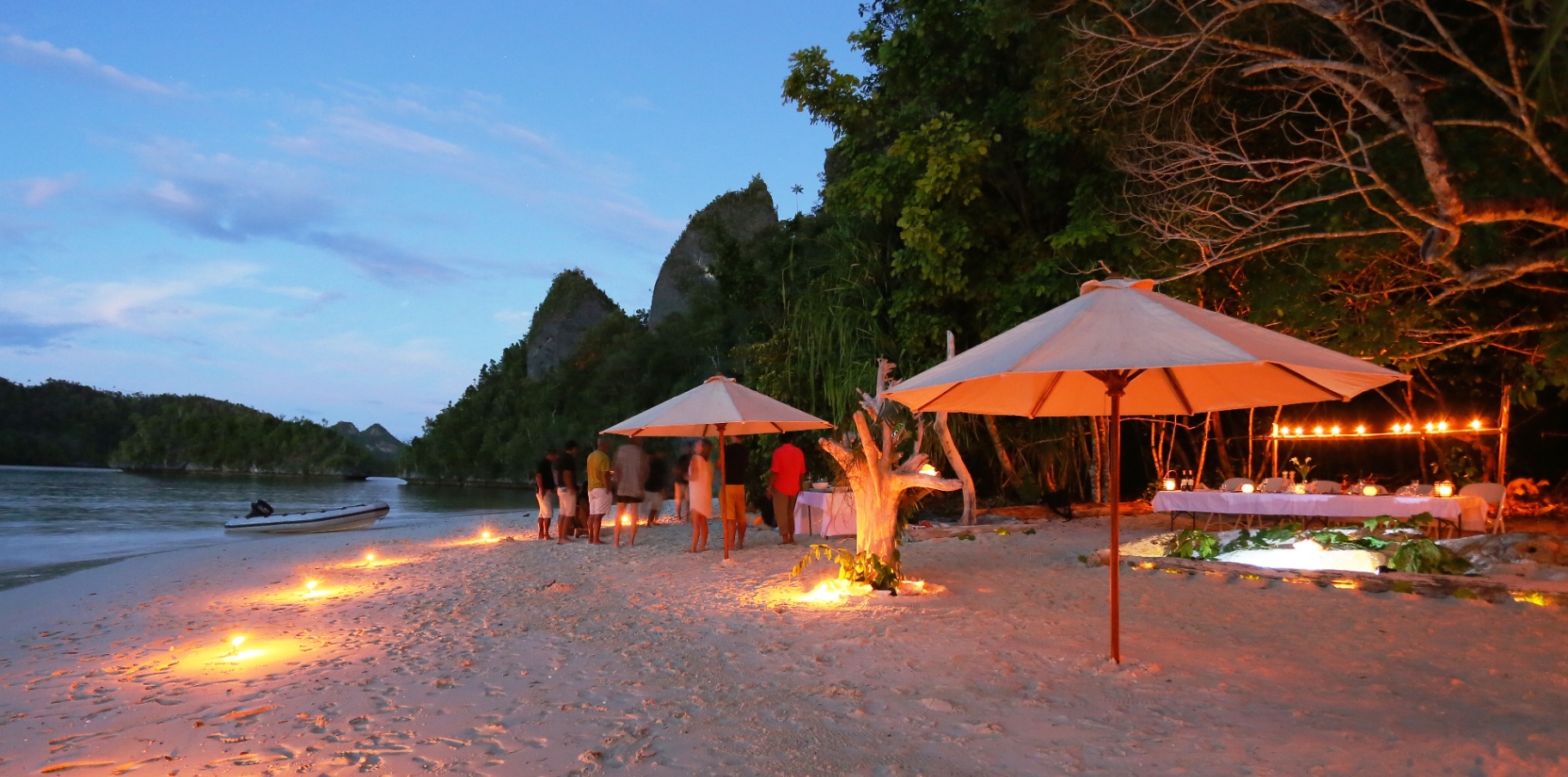 Candlelit beach dinner setup on Wayag sand at dusk with karst peaks and lantern-lit driftwood