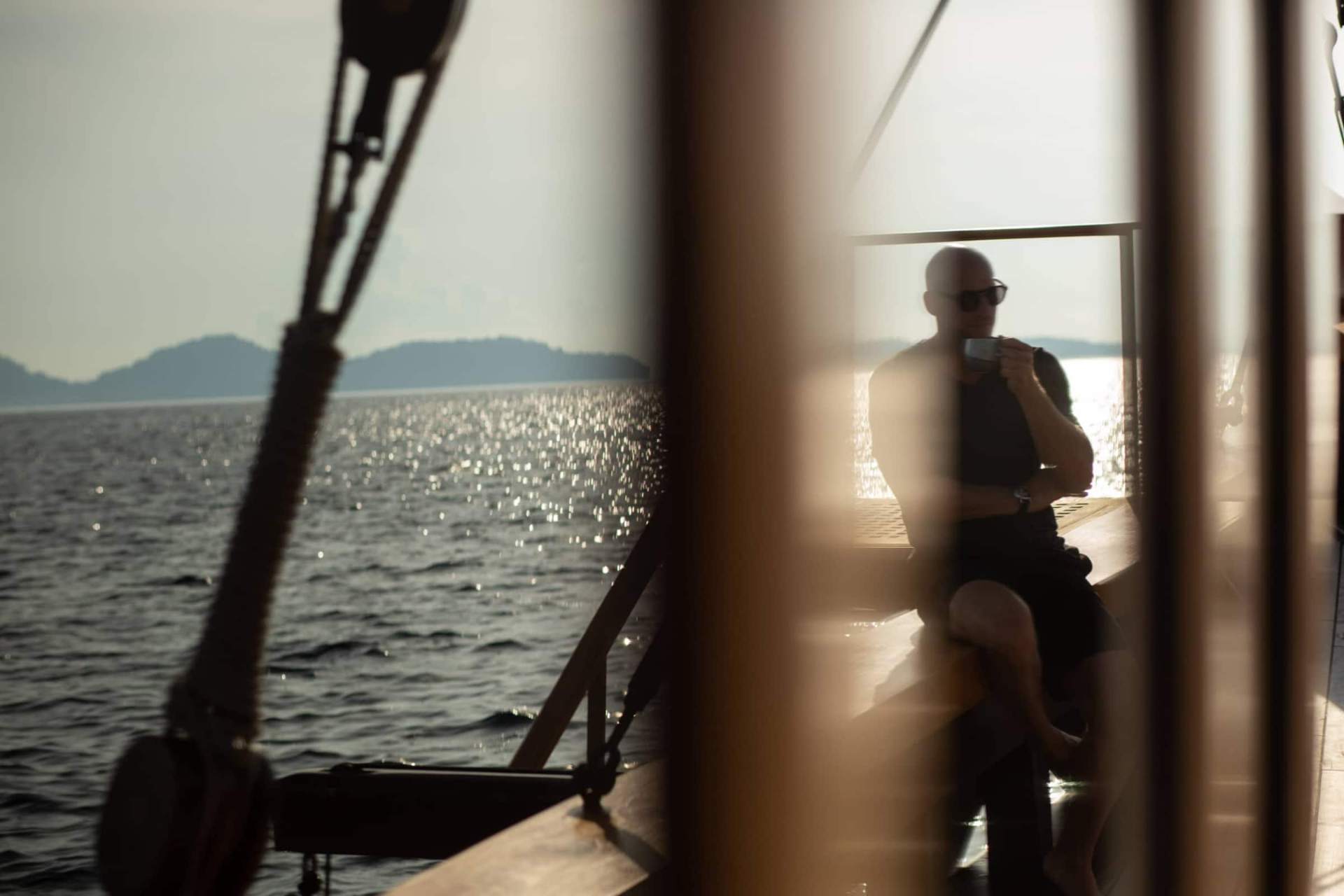 Guest reflected in Silolona's varnished timber taking morning coffee on deck at sunrise