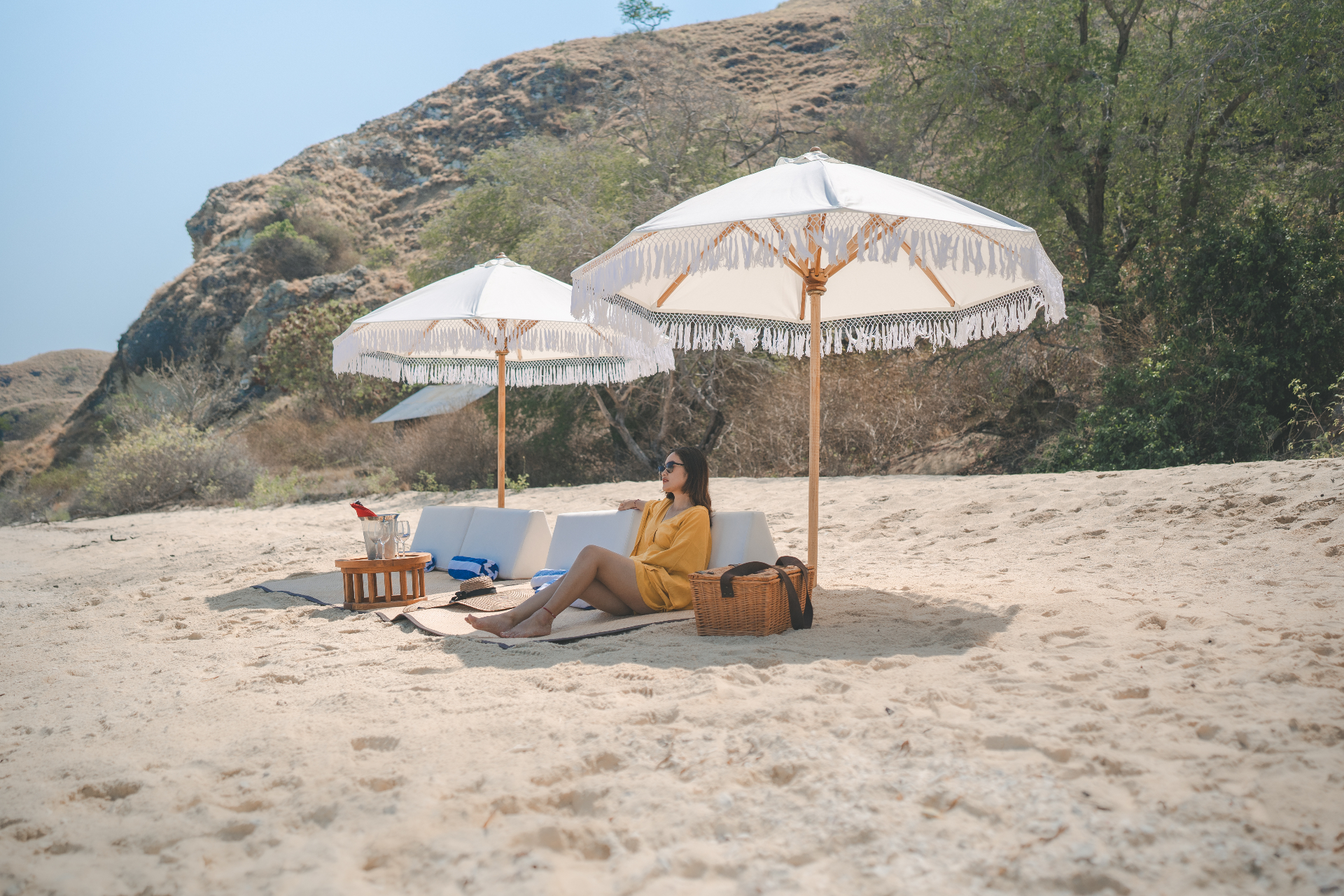 Twin fringed umbrellas shading beach loungers and a picnic hamper on a secluded Komodo cove arranged by Mischief