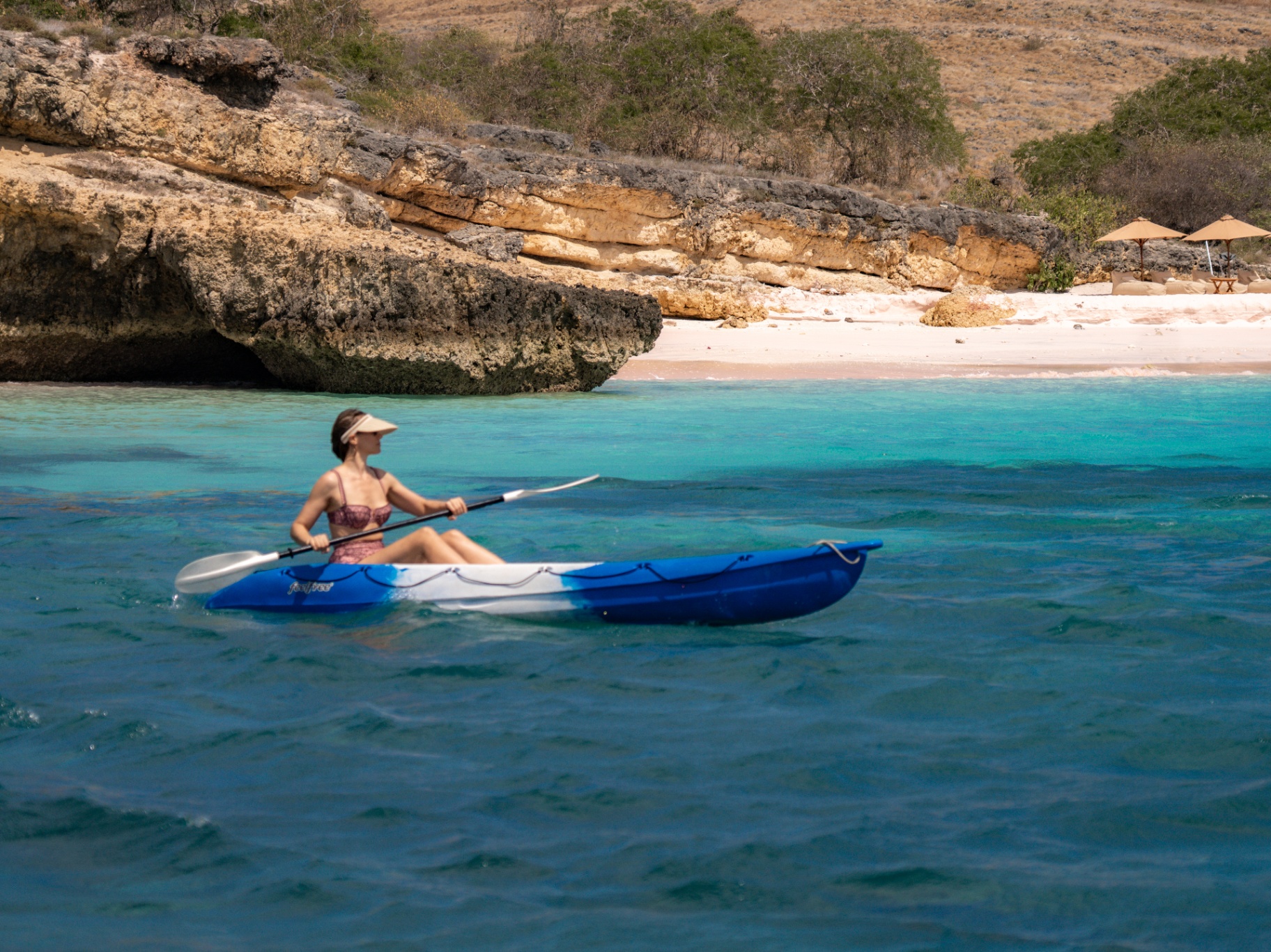 Guest kayaking in a clear turquoise cove beside a pink-sand beach with sun loungers under umbrellas