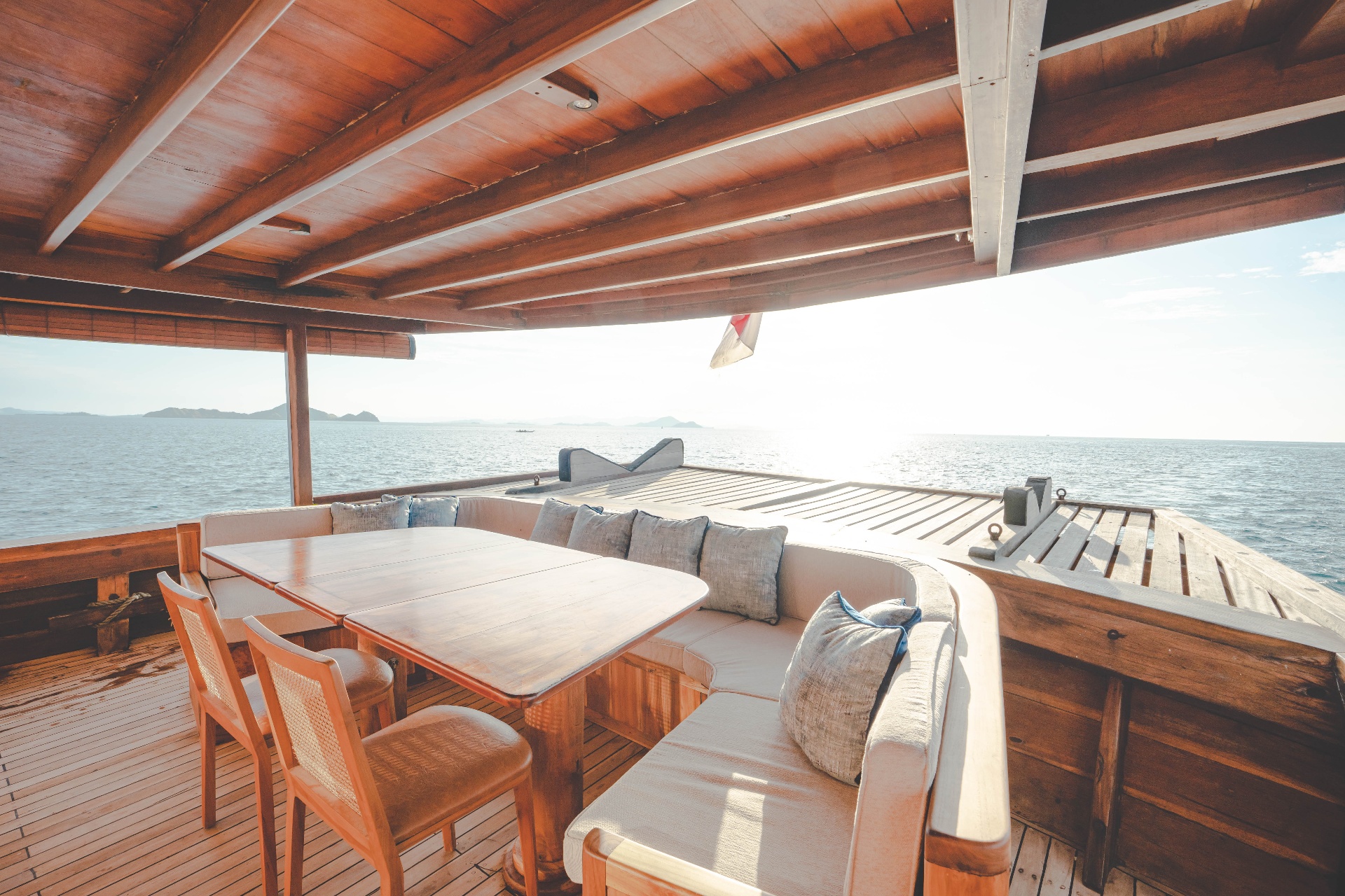 Mischief covered bow lounge with wraparound cushioned bench, teak table and cane chairs overlooking Komodo waters at sunset