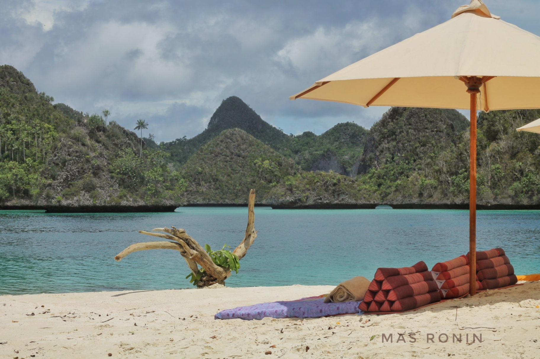 Thai triangle cushions and parasol on a white-sand beach facing turquoise water and Wayag karst peaks