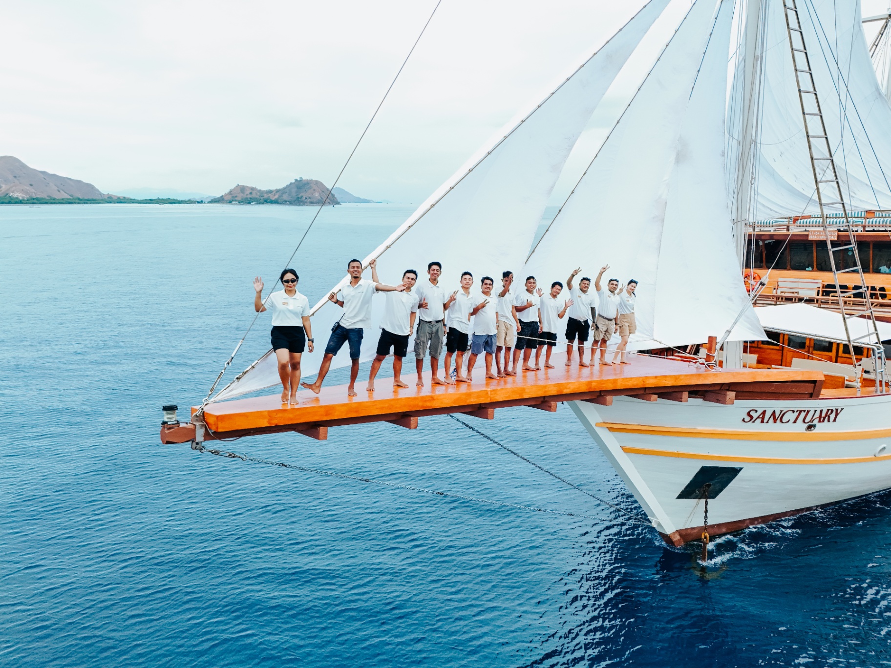 Sanctuary crew standing along the yacht's long teak bowsprit waving to camera
