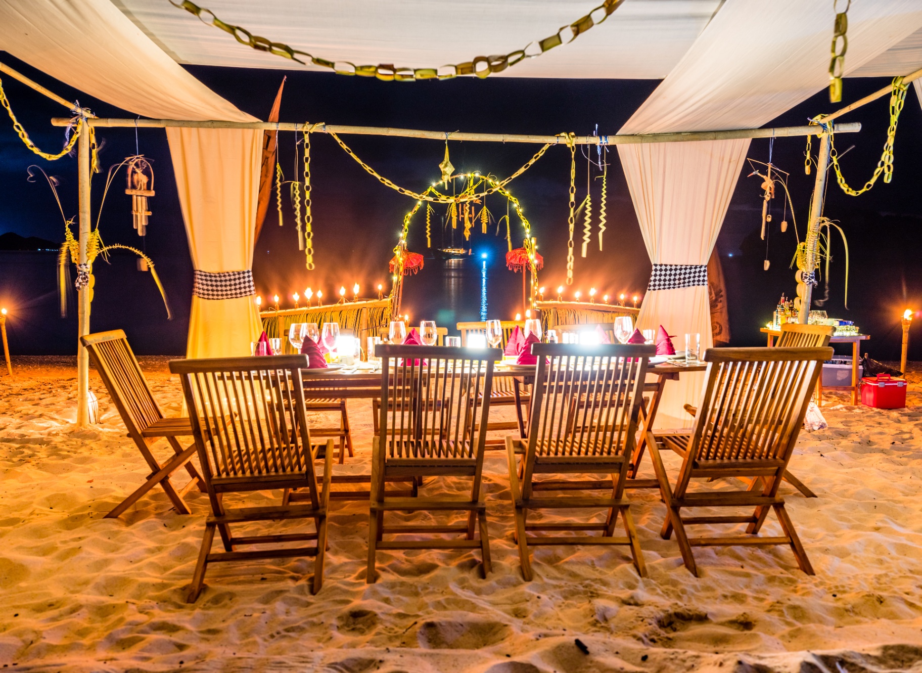 Candlelit beach dinner set under a draped canopy with lanterns and tiki torches lighting the sand at night