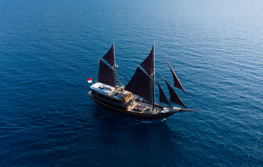 Aerial view of Dunia Baru phinisi under full black sails in the deep-blue waters of Komodo National Park
