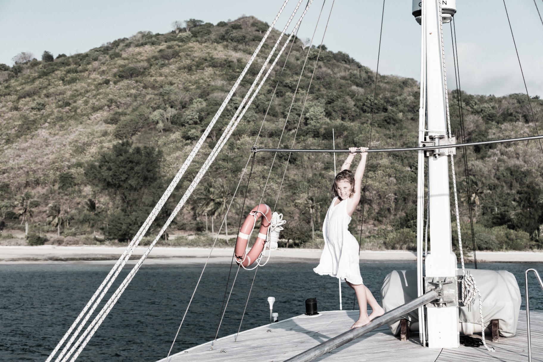 Child playing on Splendour's bow beside the mast and life ring with a beach and hillside behind