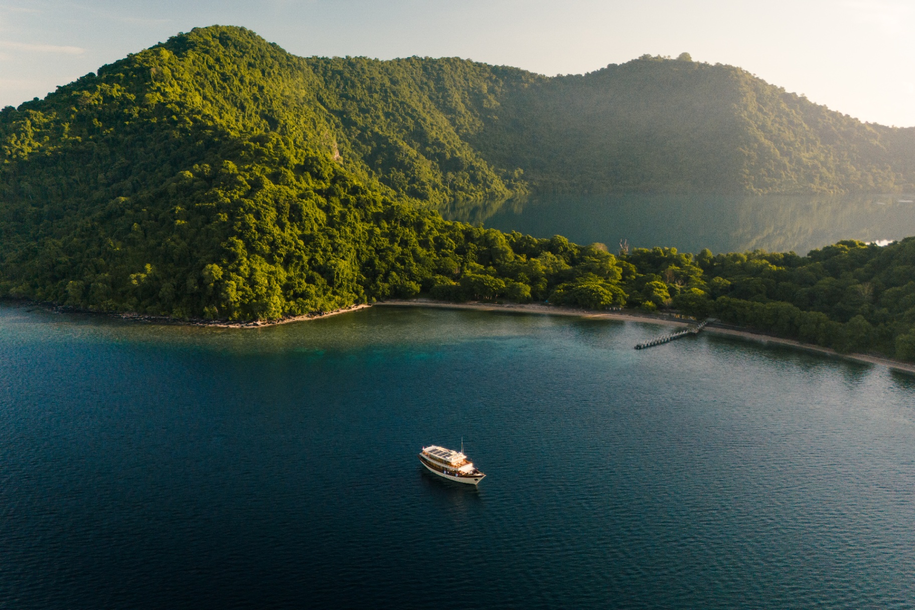 Mischief off a rainforest-clad shoreline with a long wooden jetty and Sunda Islands beyond