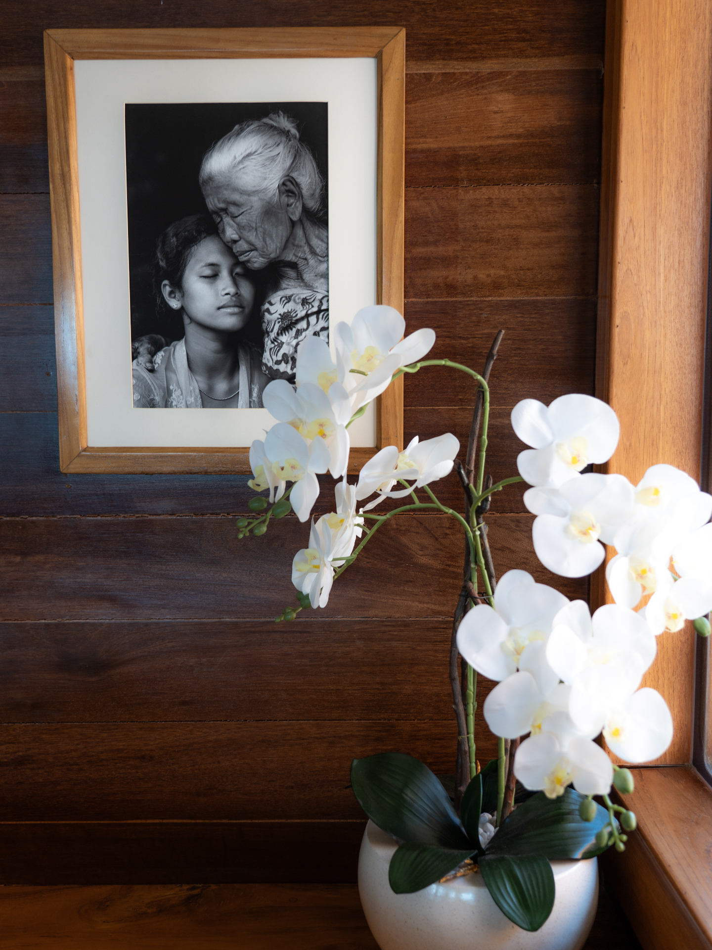Framed black-and-white portrait of an Indonesian grandmother and granddaughter beside white orchids in a Lamima interior