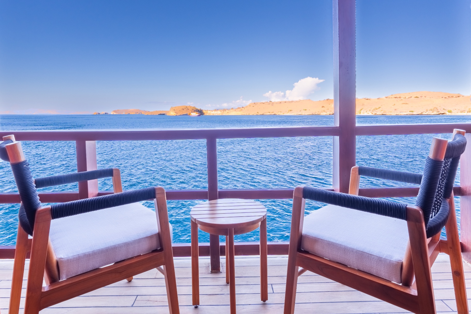 Private balcony on Sanctuary's deluxe cabin with two teak armchairs and side table overlooking the sea and dry hills