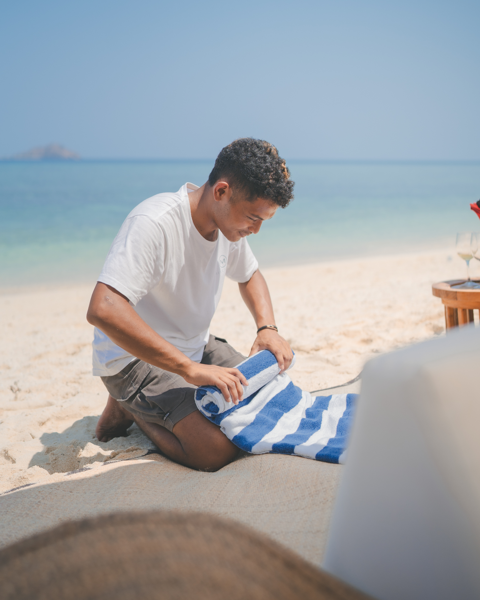 Mischief crew member rolling striped beach towels on the sand while setting up a shoreline picnic in Komodo