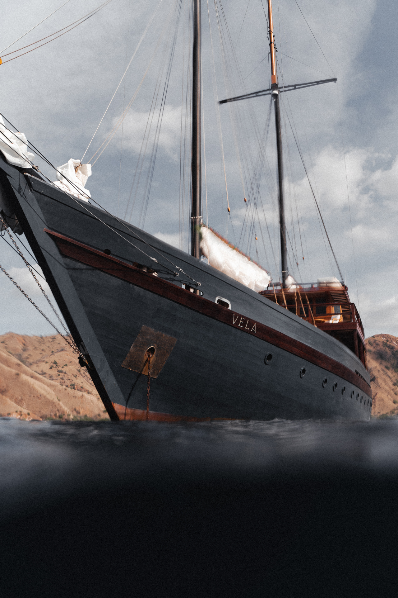 Water-level view of Vela's dark hull and bow with 'VELA' name plate and rigging above