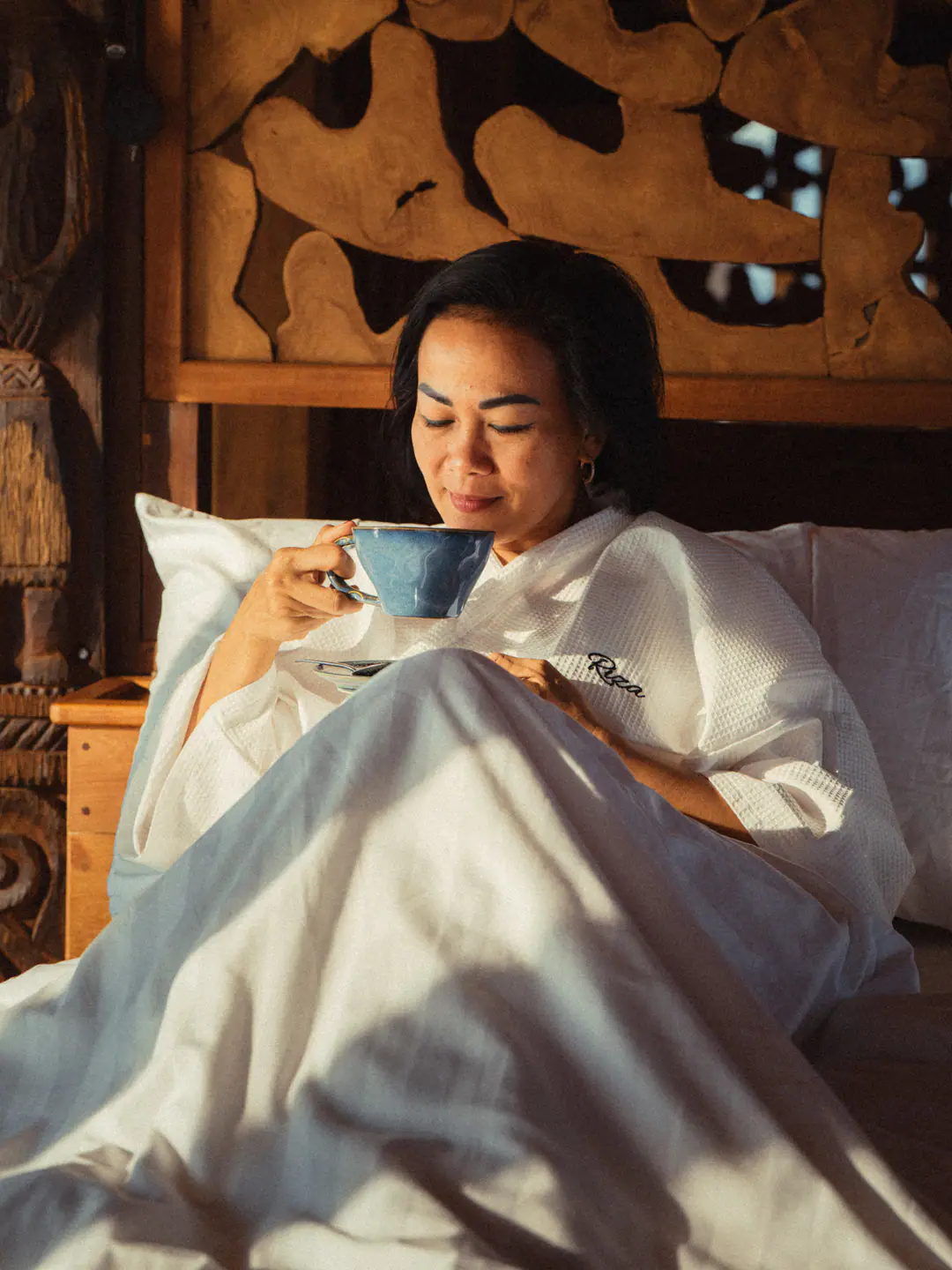 Guest in white robe savouring morning coffee in bed on The Oracle with carved wooden headboard