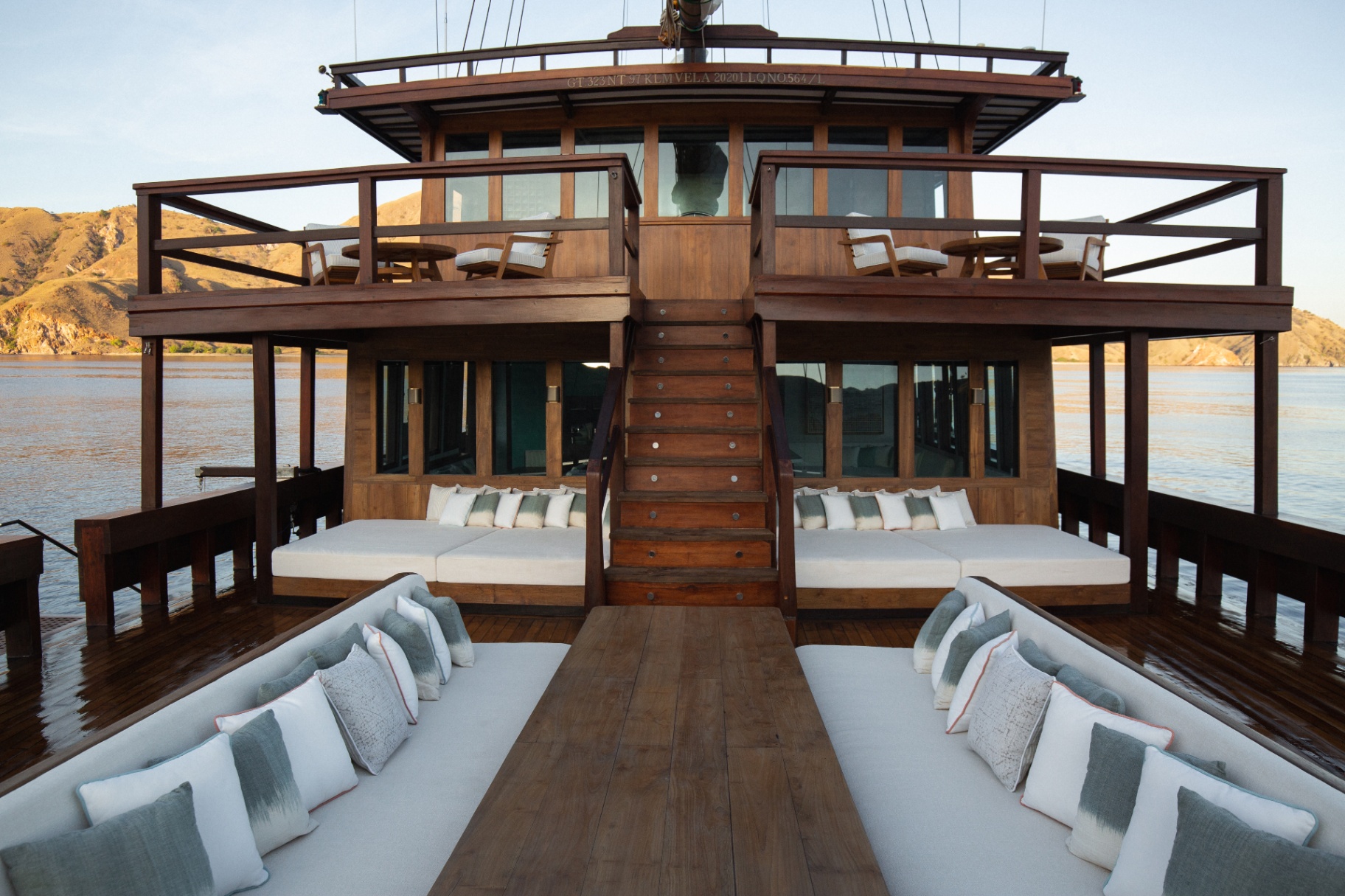 Symmetrical view down Vela's aft deck with facing daybeds, sunloungers and stairs up to the bridge deck against Komodo hills