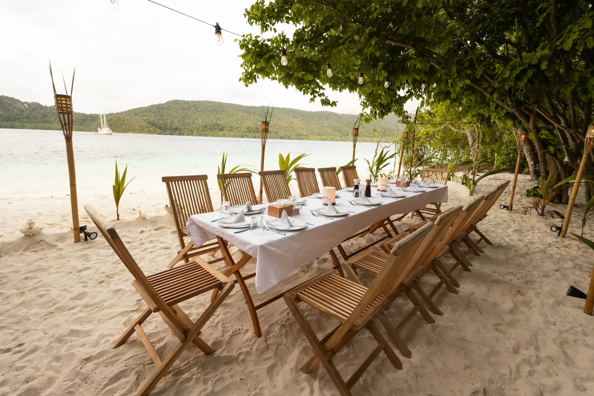 Private beach dinner set for Mutiara Laut guests with tiki torches and yacht anchored offshore