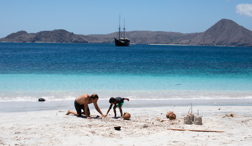 Father and child building sandcastles on a white-sand beach with Dunia Baru anchored offshore