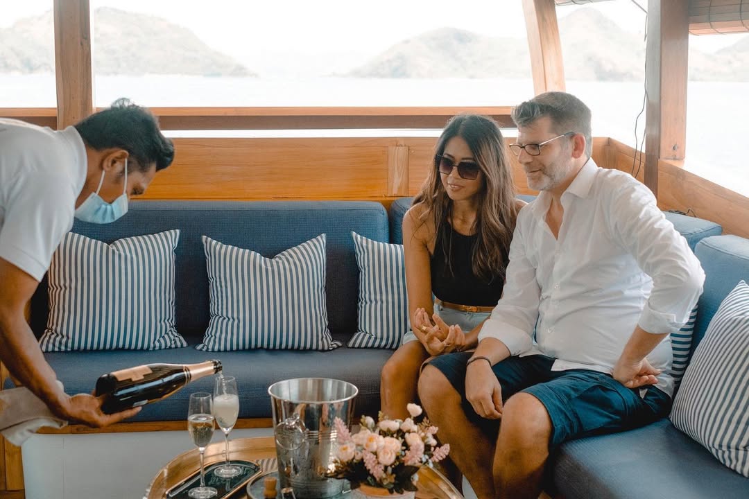 Couple seated on Samara II's blue-and-white striped saloon sofa while a crew member pours champagne
