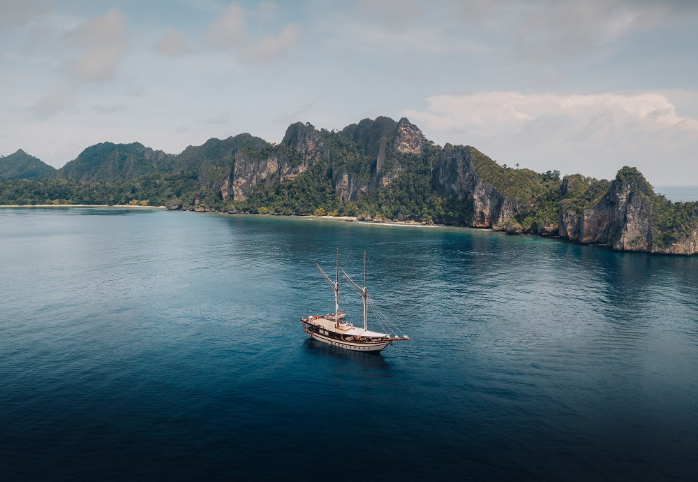 Aerial of Senja at anchor in a deep blue bay ringed by karst cliffs in Raja Ampat