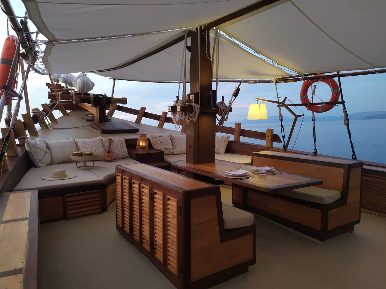 Foredeck of Senja at dusk with lit lamps, cushioned lounge and dining table set for service