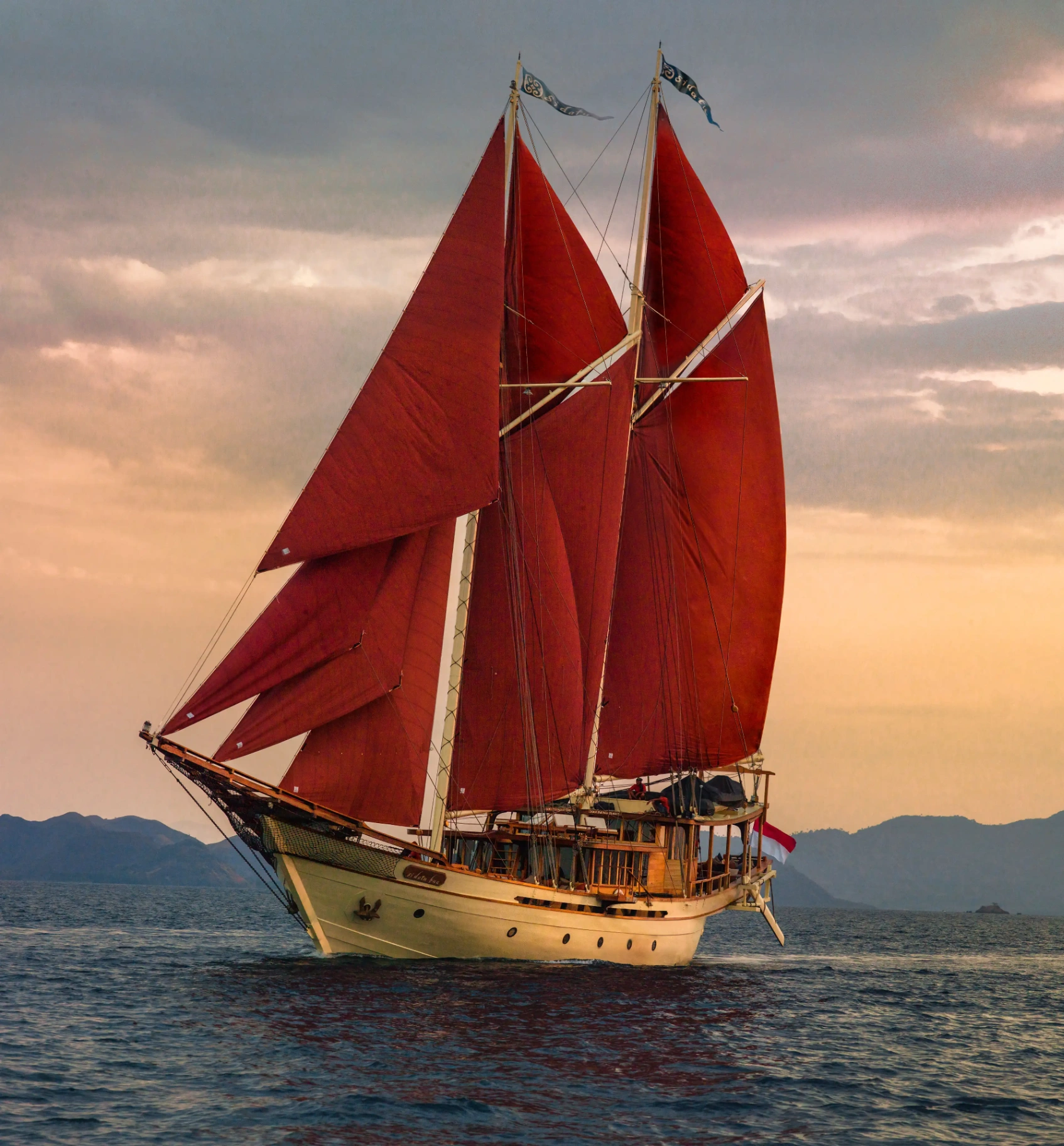 Si Datu Bua luxury phinisi under full crimson gaff-rigged sails at golden hour in Komodo