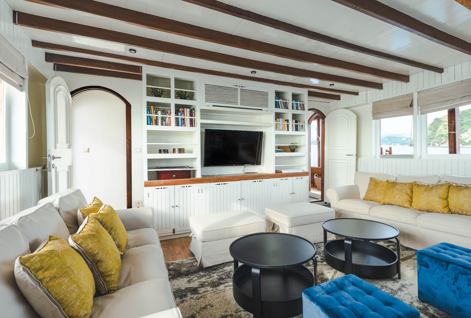 Mischief indoor saloon lounge with built-in bookshelves, TV, white sofas and blue velvet ottomans