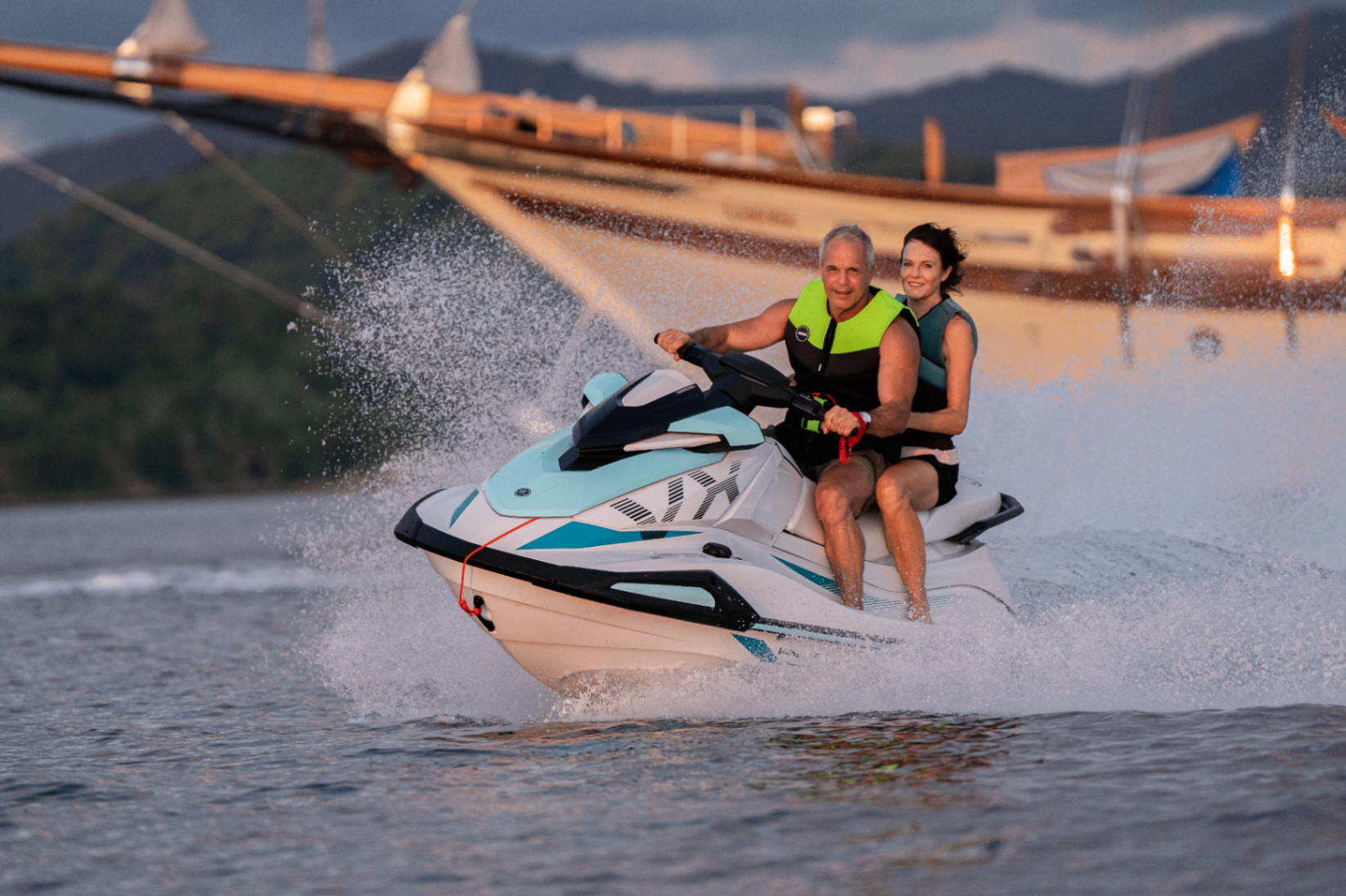 Couple riding a jetski in golden evening light with Lamima's bow and rigging blurred behind them
