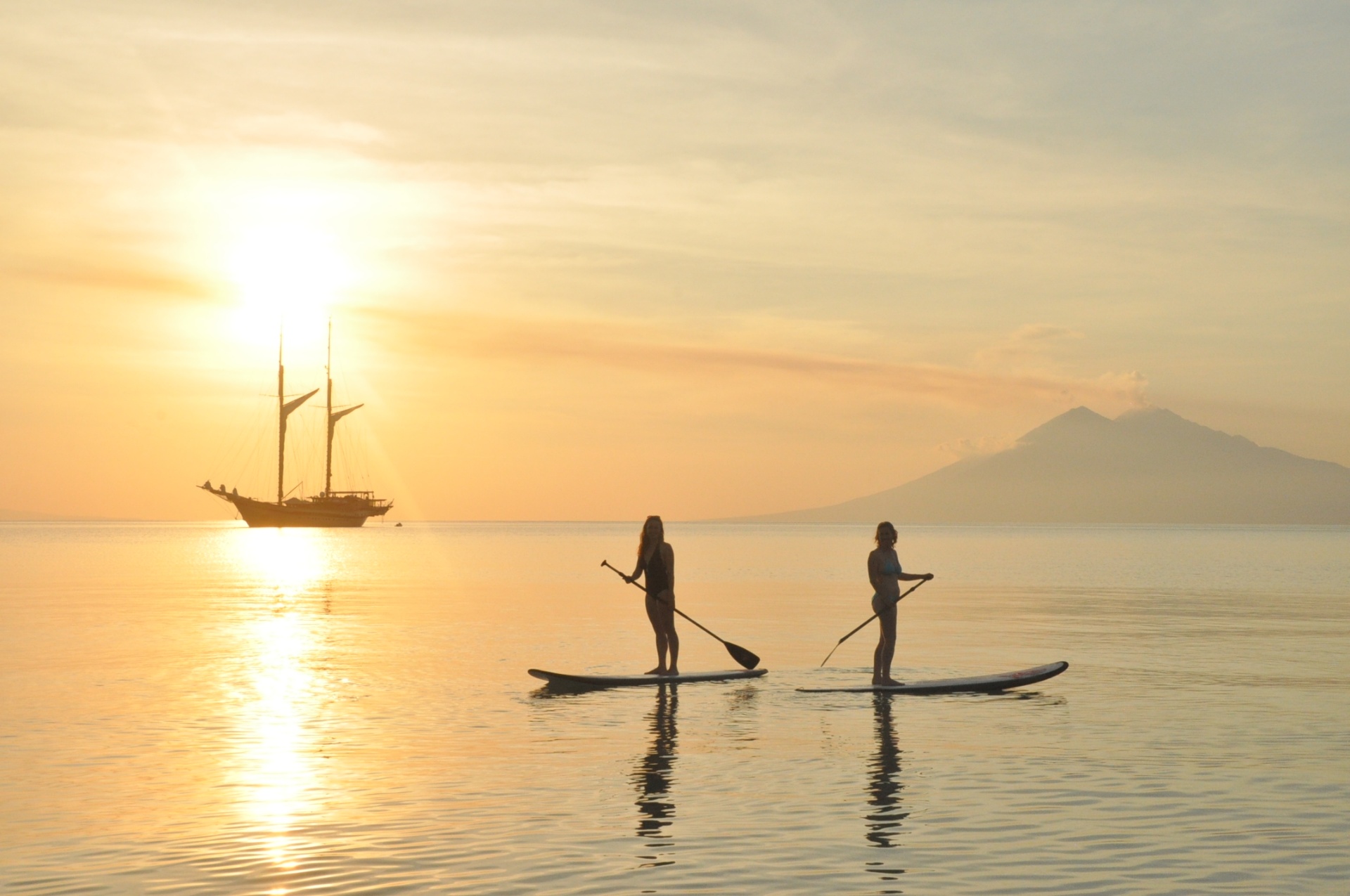 Two guests stand-up paddleboarding on a mirror-calm sea at sunset with Lamima and a distant volcano behind them