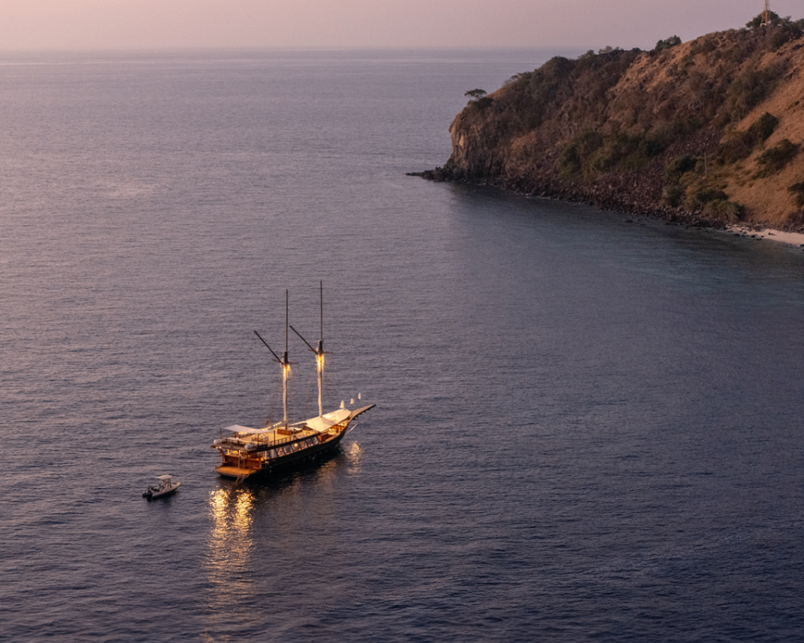 Wide aerial of Sanya glowing at anchor in evening light near a rocky island